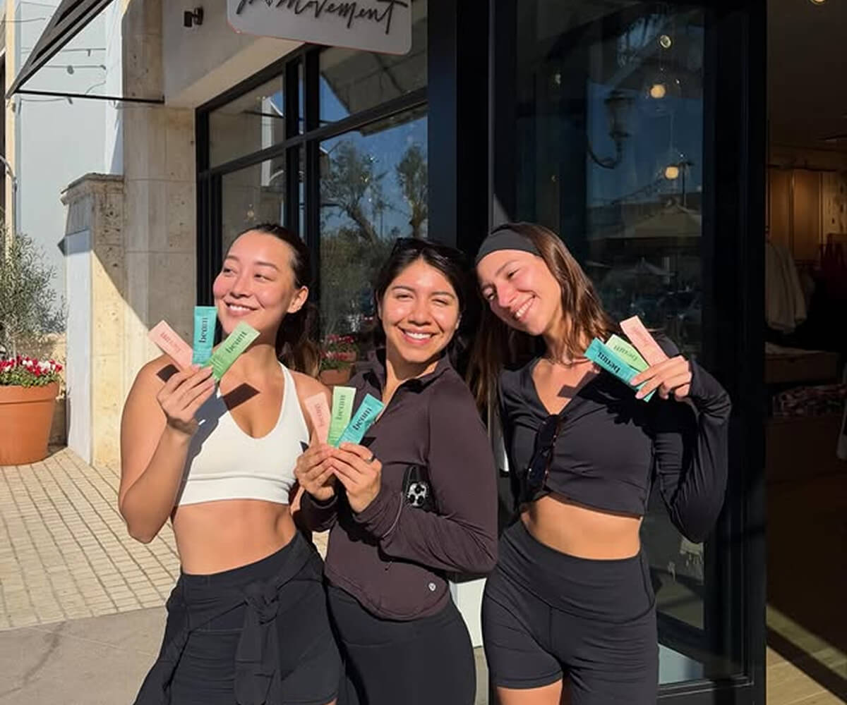Three women in athletic wear smiling and holding colorful supplement packets outside a storefront.