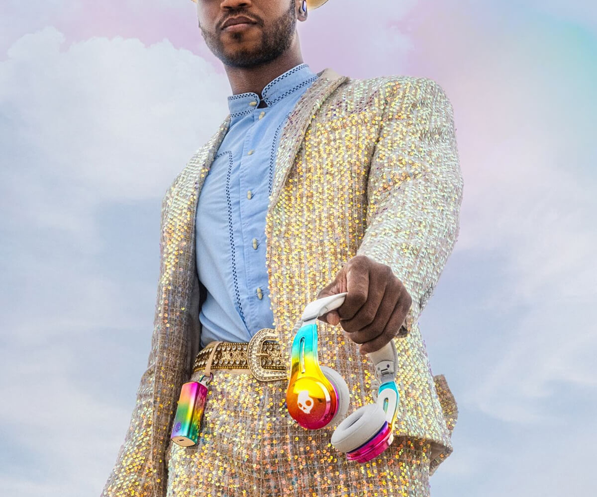 Person wearing a sequined suit holding rainbow-colored Skullcandy headphones against a cloudy sky.
