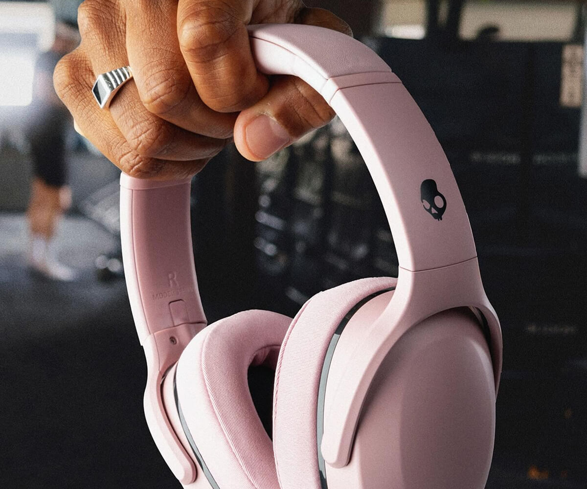 Hand holding pink over-ear headphones with a skull logo on the headband in a gym setting.