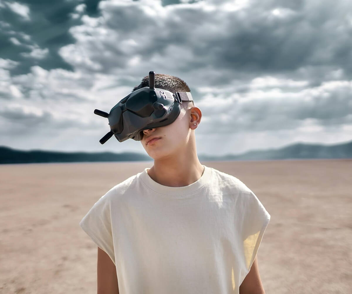 Young person outdoors wearing a white sleeveless shirt and VR goggles with antennas against a cloudy sky background.