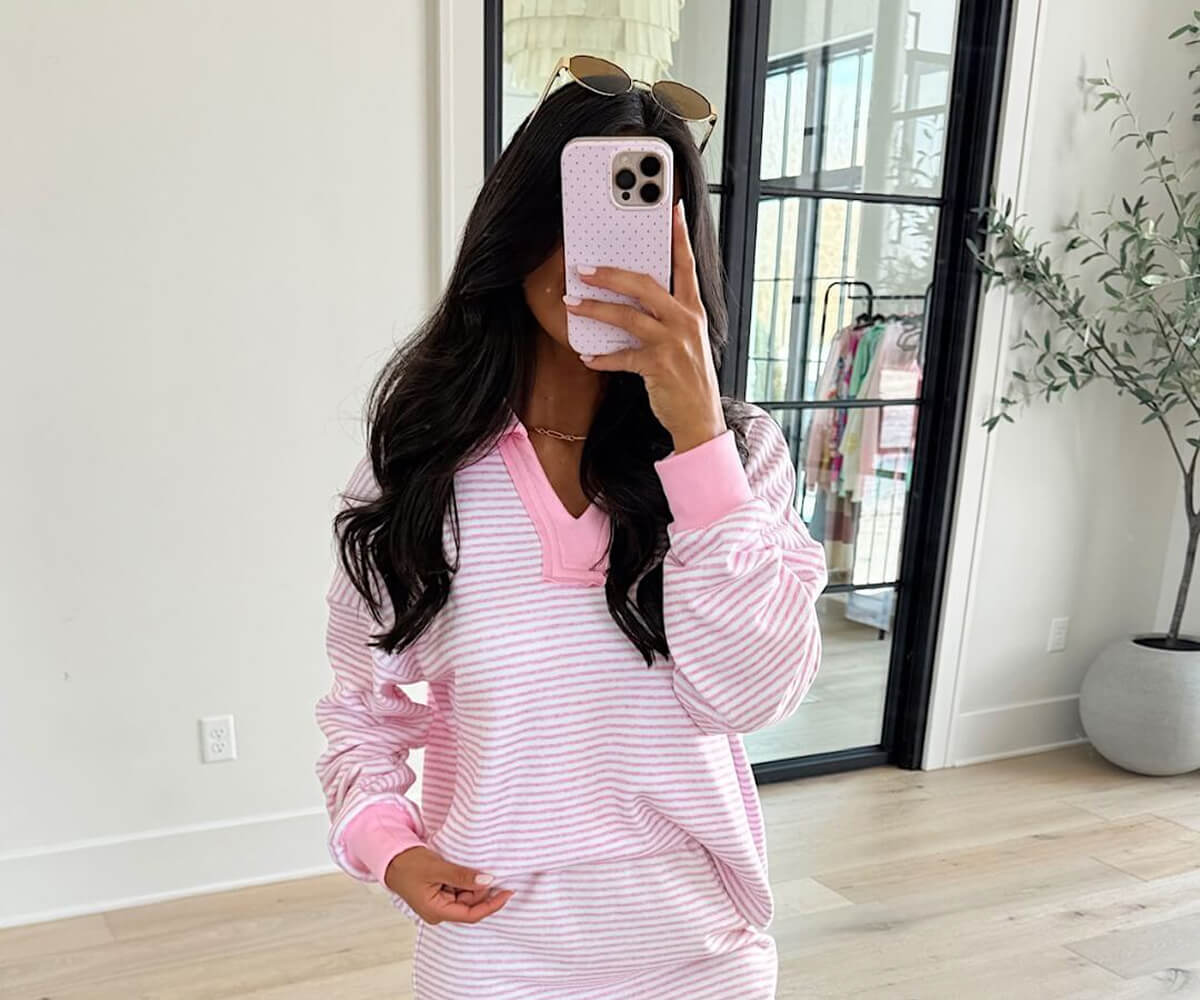 Woman wearing pink and white striped long-sleeve top taking a mirror selfie with phone covering her face.