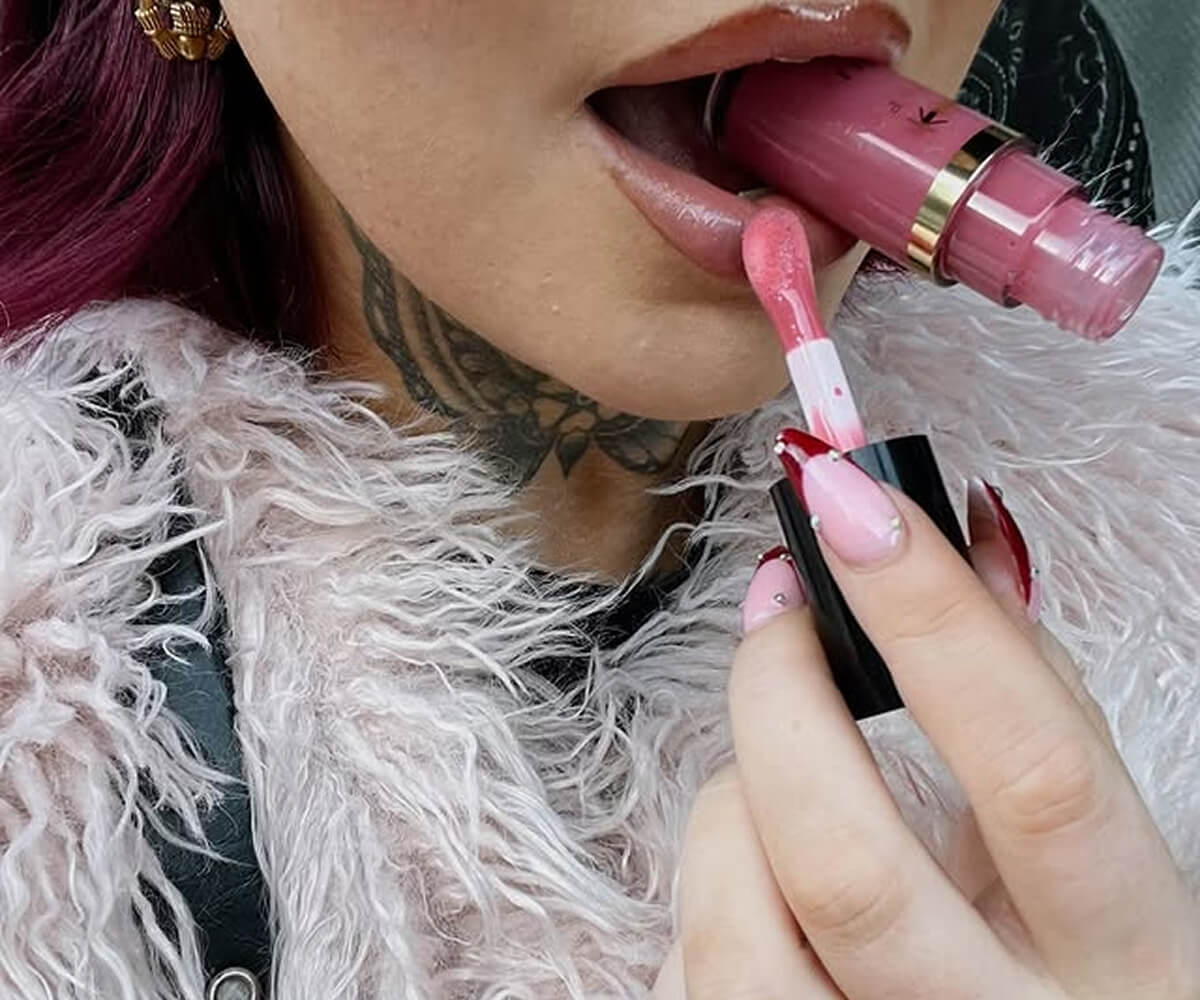 Close-up of a person with a neck tattoo and pink fur jacket applying pink lip gloss with the applicator while holding the lip gloss tube in their mouth.