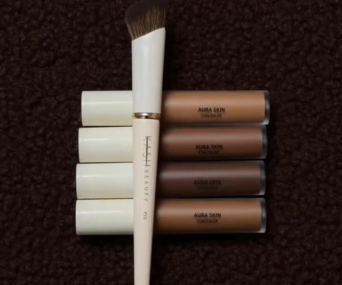 Makeup brush marked KASH BEAUTY and four shades of AURA SKIN concealer tubes arranged on a dark textured background.