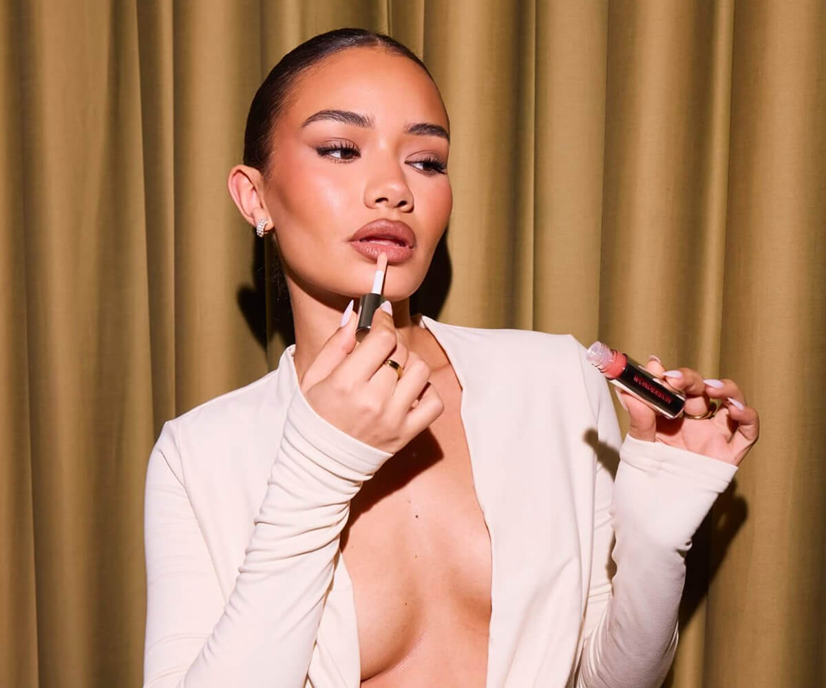 Woman in a white top applying nude lip gloss in front of a gold curtain.