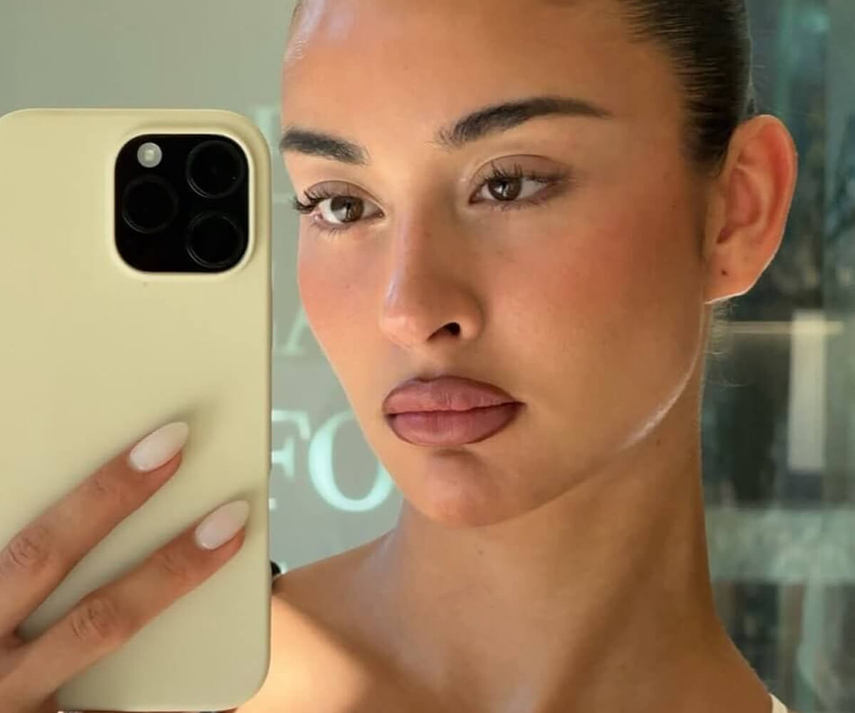 Close-up portrait of a woman with long wavy brown hair and natural makeup looking at the camera indoors.