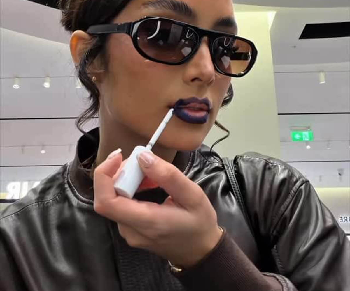Person wearing black sunglasses and a leather jacket applying dark purple lipstick indoors.
