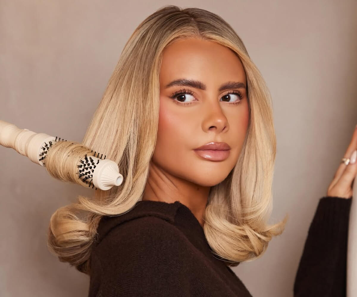 Blonde woman with styled hair holding a round hairbrush curling the ends of her hair.