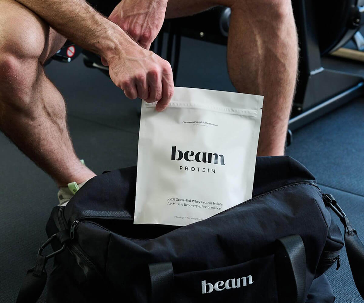 Person placing a white packet of Beam Protein powder into a black gym bag with the brand name 'beam' on it.