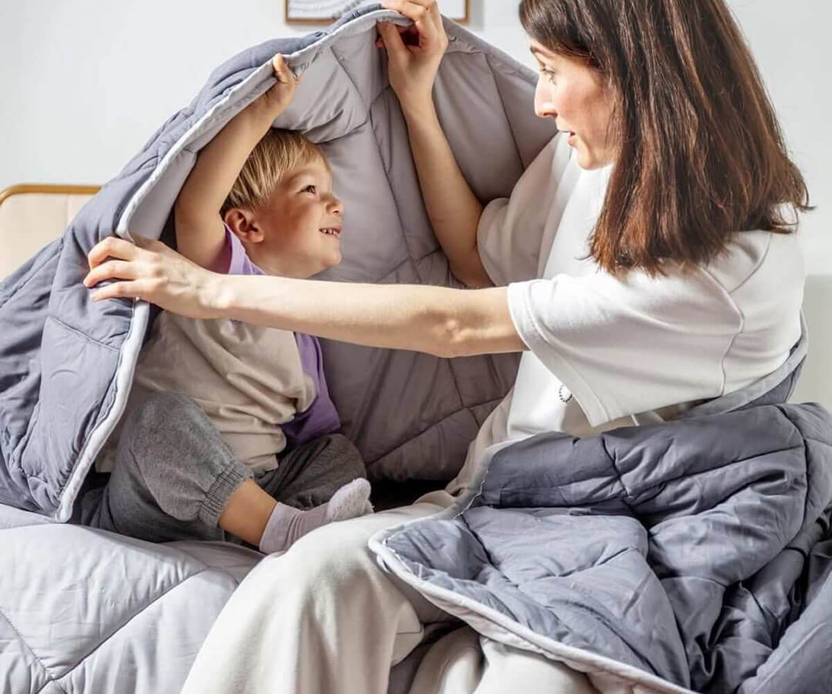 Mother and young child playing under a gray blanket on a bed, smiling at each other.