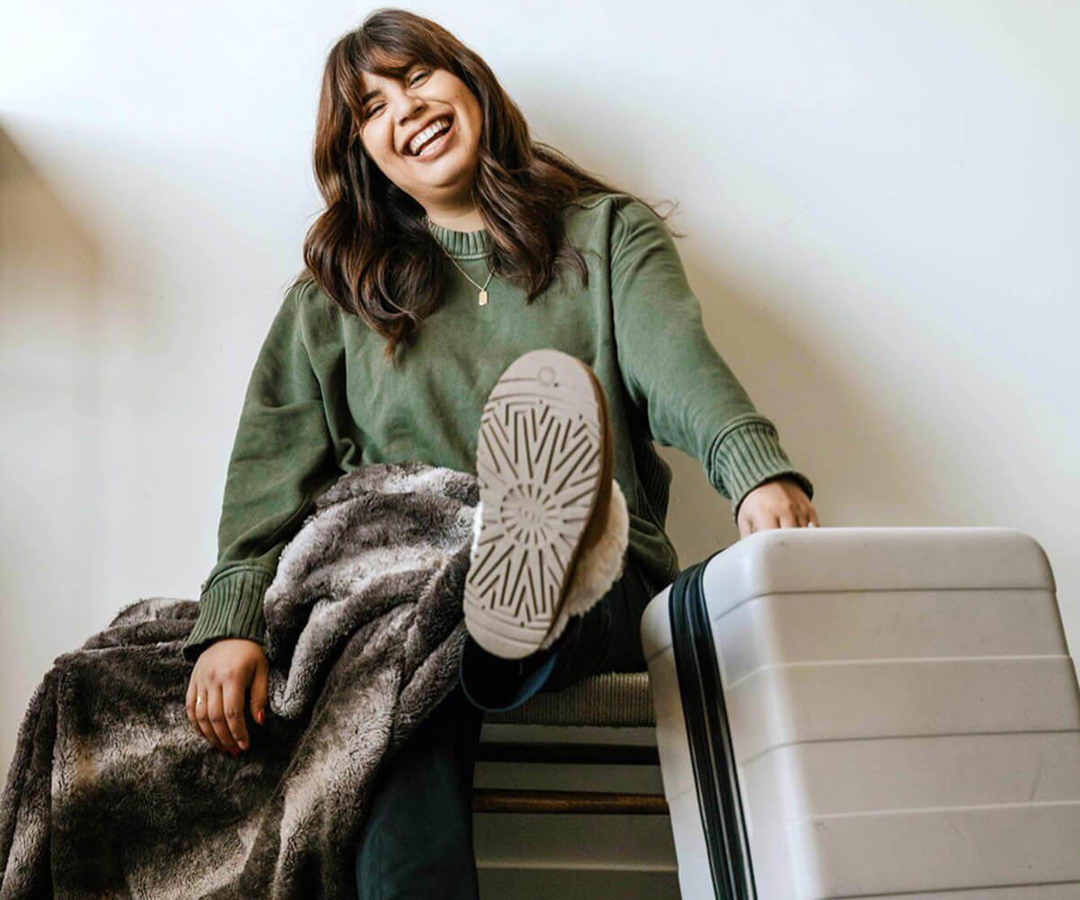 Smiling woman in green sweater sitting with a furry blanket and a large white suitcase.
