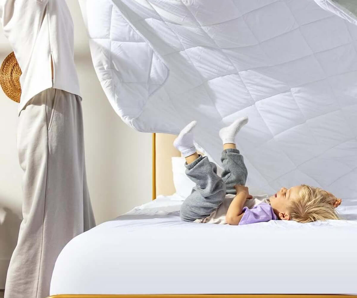 Child lying on a bed kicking legs up while an adult is making the bed with a white quilt.