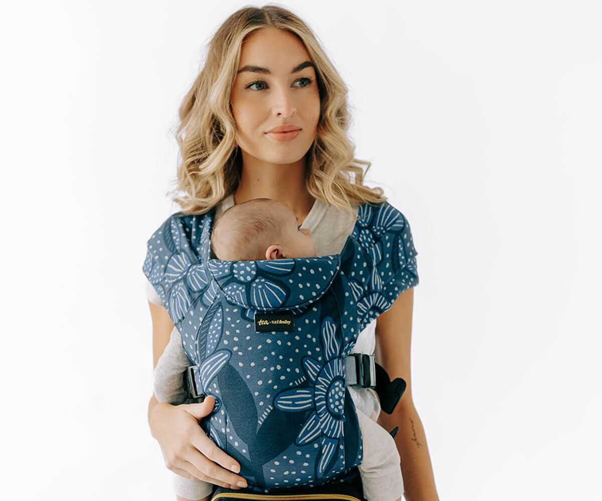 Blonde woman carrying a sleeping baby in a blue floral-patterned baby carrier.