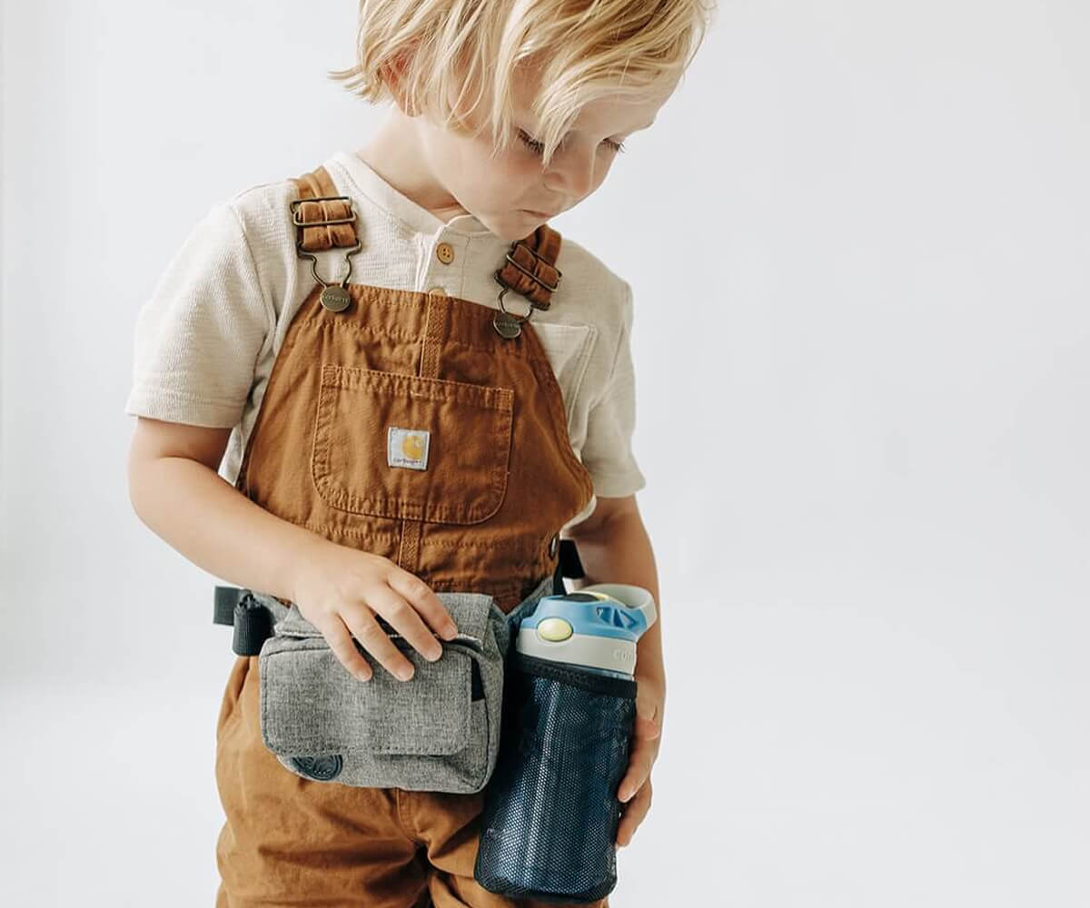 Young child with blonde hair wearing brown overalls and a white shirt, holding a gray pouch and a blue water bottle.