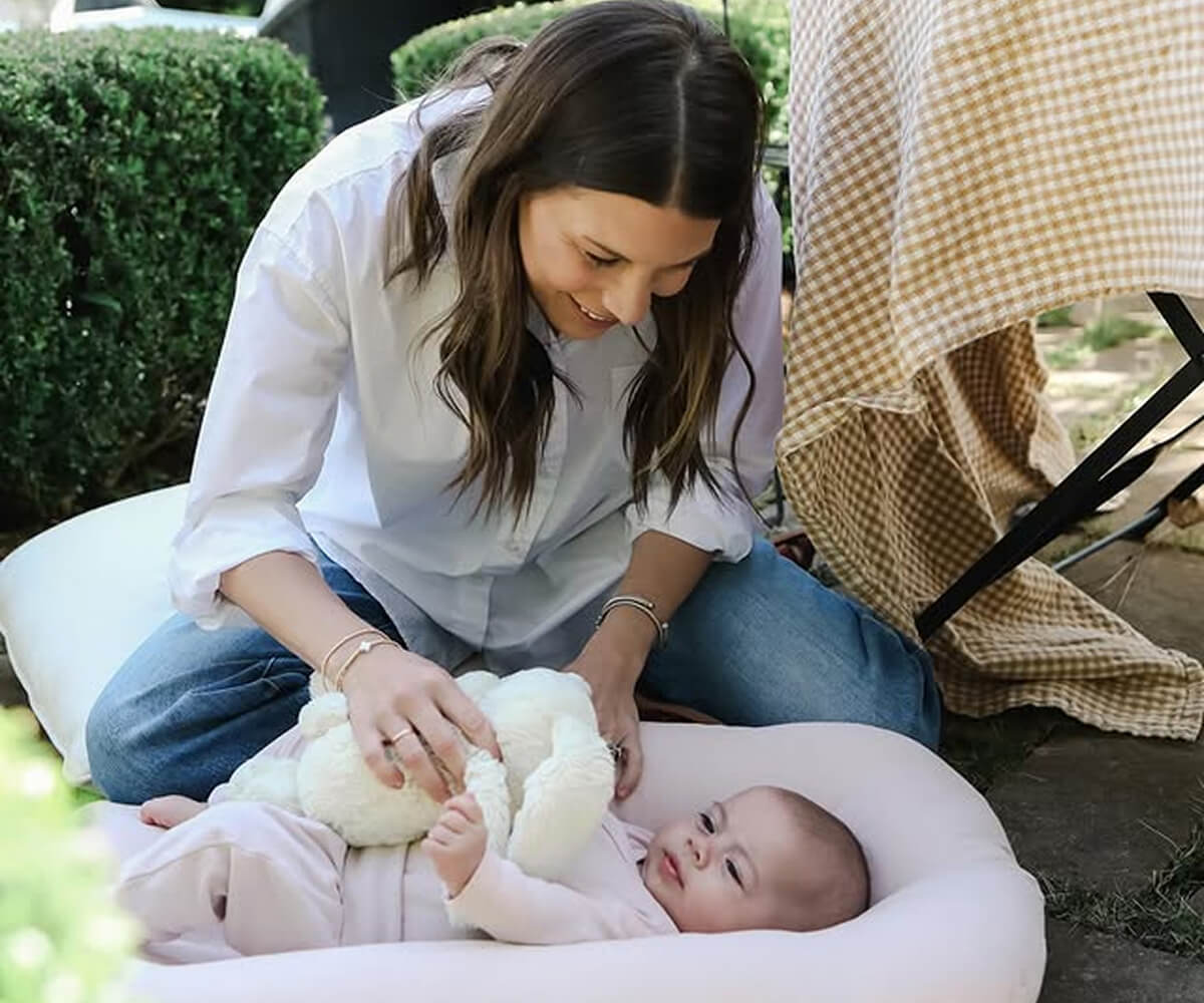 A woman with brown hair smiles while playing with a baby lying on a pink cushion, holding a white plush toy outdoors.