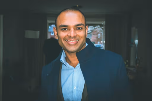 Smiling man wearing a blue shirt and dark jacket standing indoors with a blurred background.
