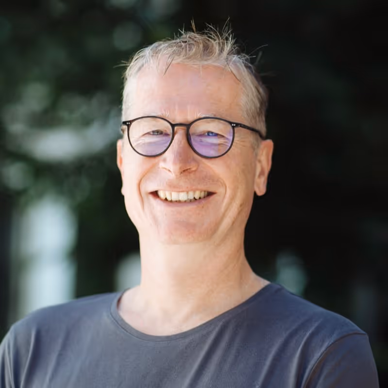 Smiling middle-aged man with short blond hair and glasses wearing a dark gray t-shirt standing outdoors with blurred greenery in the background.
