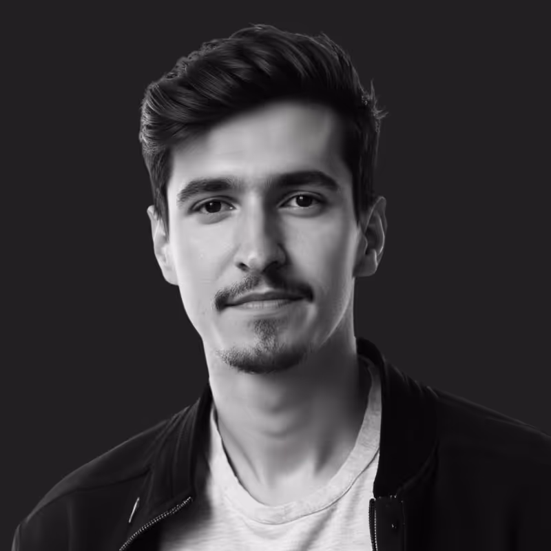 Black and white portrait of a young man with short hair, light facial hair, and a slight smile, wearing a jacket over a t-shirt against a dark background.