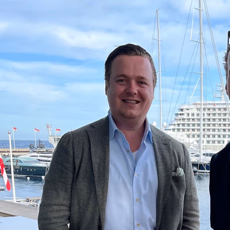 Smiling man in a gray blazer and light blue shirt standing in front of a marina with yachts and a cruise ship under a partly cloudy sky.
