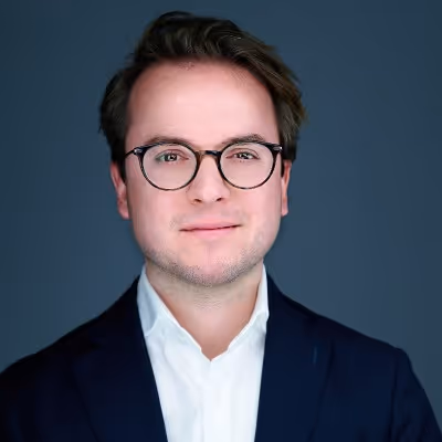 Young man wearing black round glasses, a white shirt, and a dark blazer against a dark blue background.