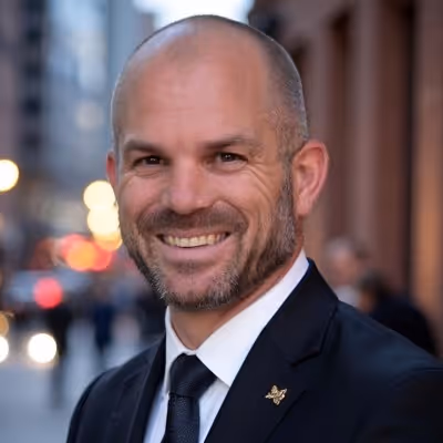 Smiling bald man with a short beard wearing a black suit and tie in an urban street setting.