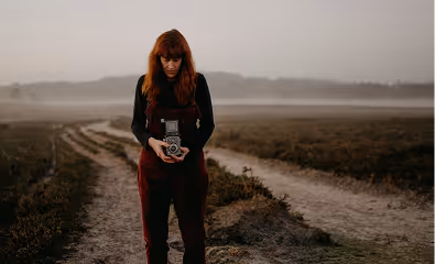 Woman with red hair standing on a dirt path in a misty field holding a vintage camera.