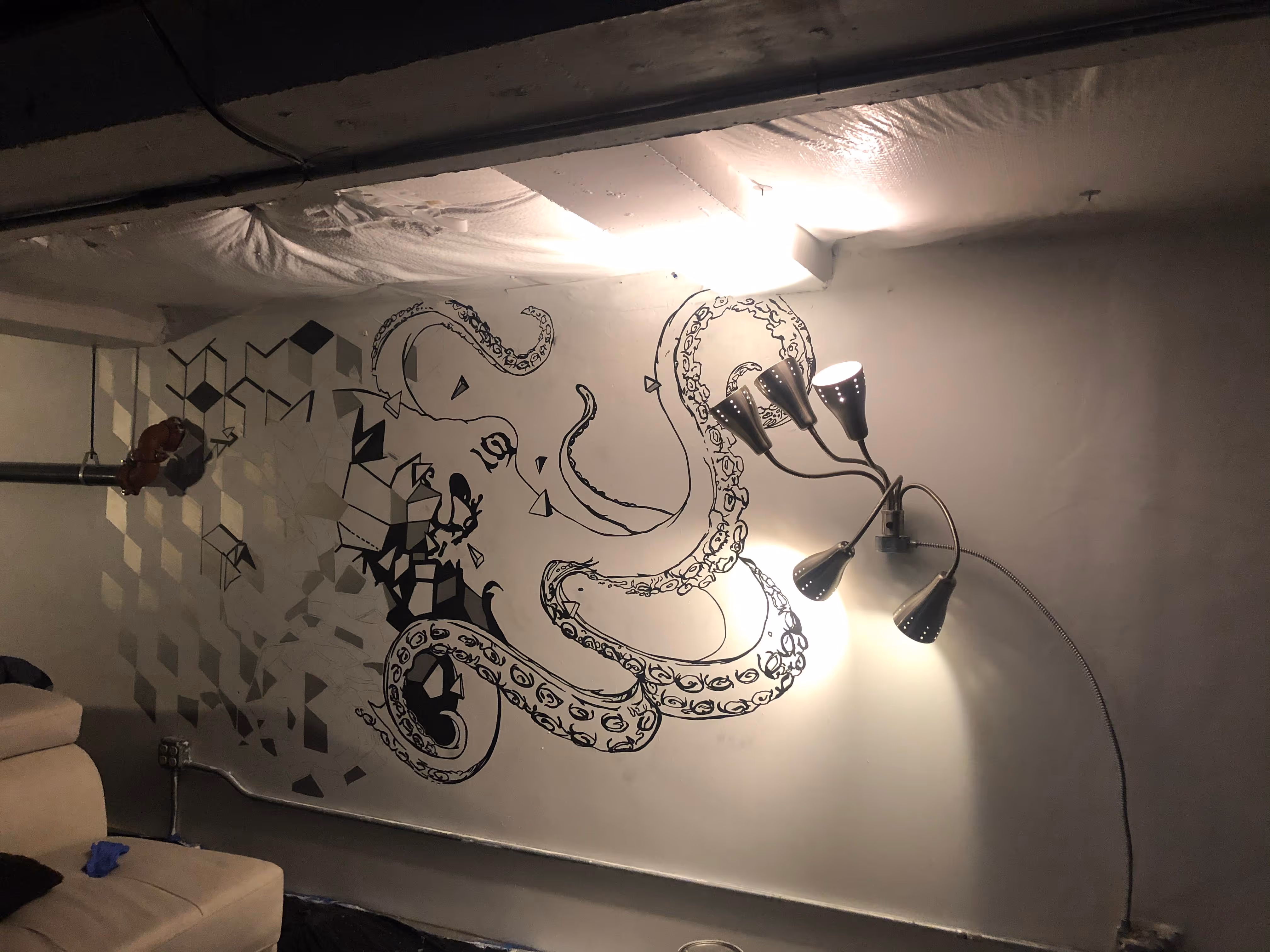 Wall mural of an octopus with geometric shapes on a basement wall next to a beige couch and a multi-headed lamp.