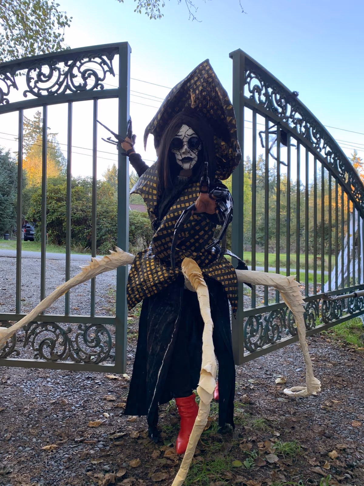 Person in elaborate Halloween costume with cracked white mask, large hat, mechanical claw gloves, red boots, and skeletal appendages, standing in front of open green wrought iron gates outdoors.