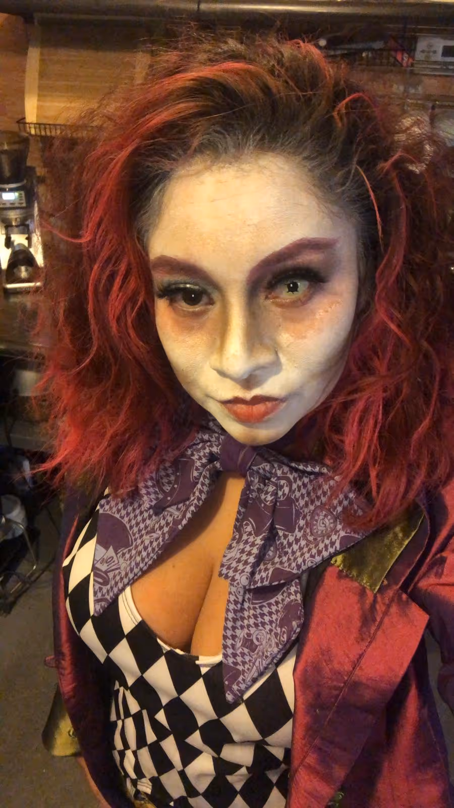 Person with red and black curly hair wearing dramatic pale face makeup, purple patterned scarf, purple and gold jacket, and black-and-white checkered outfit.