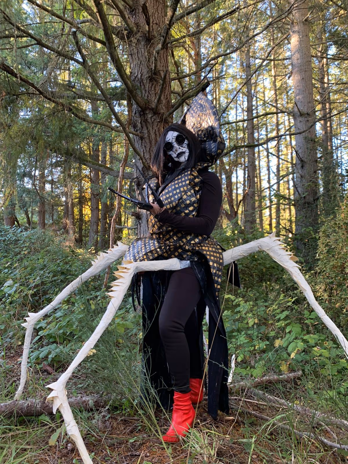 Person dressed in a spider-inspired costume with a checkered gold and black body, large spider legs, face painted white with black spider details, and red boots standing in a forest.