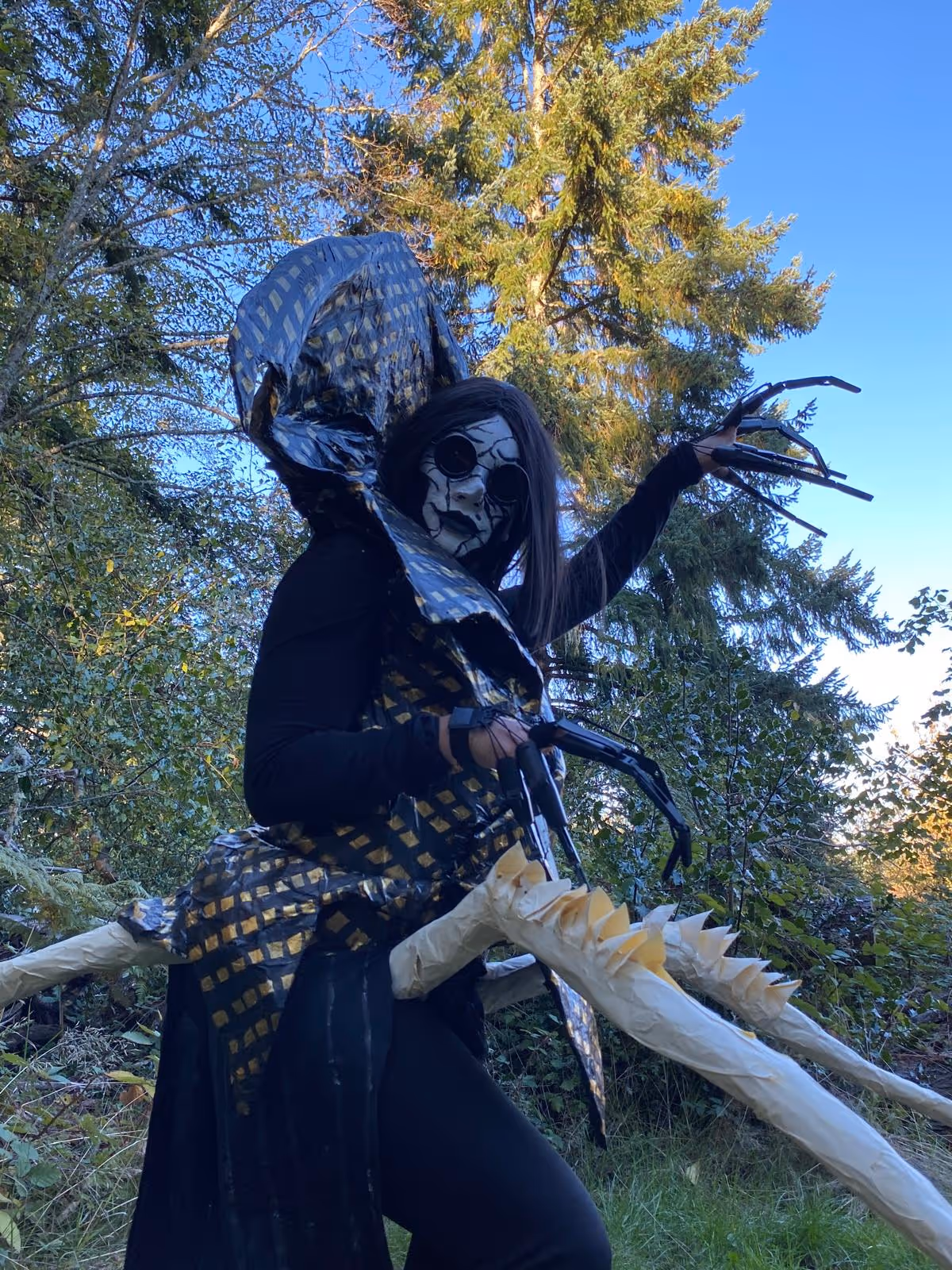 Person in a black and gold spiked costume with cracked face paint and long dark hair, posing outdoors with trees in the background.
