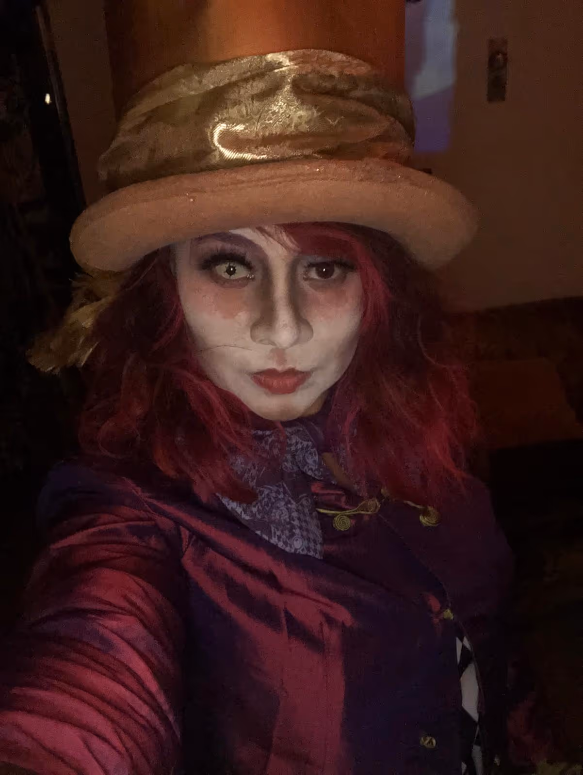 Person with red hair wearing white face makeup, a beige hat with a ribbon, and a purple jacket.