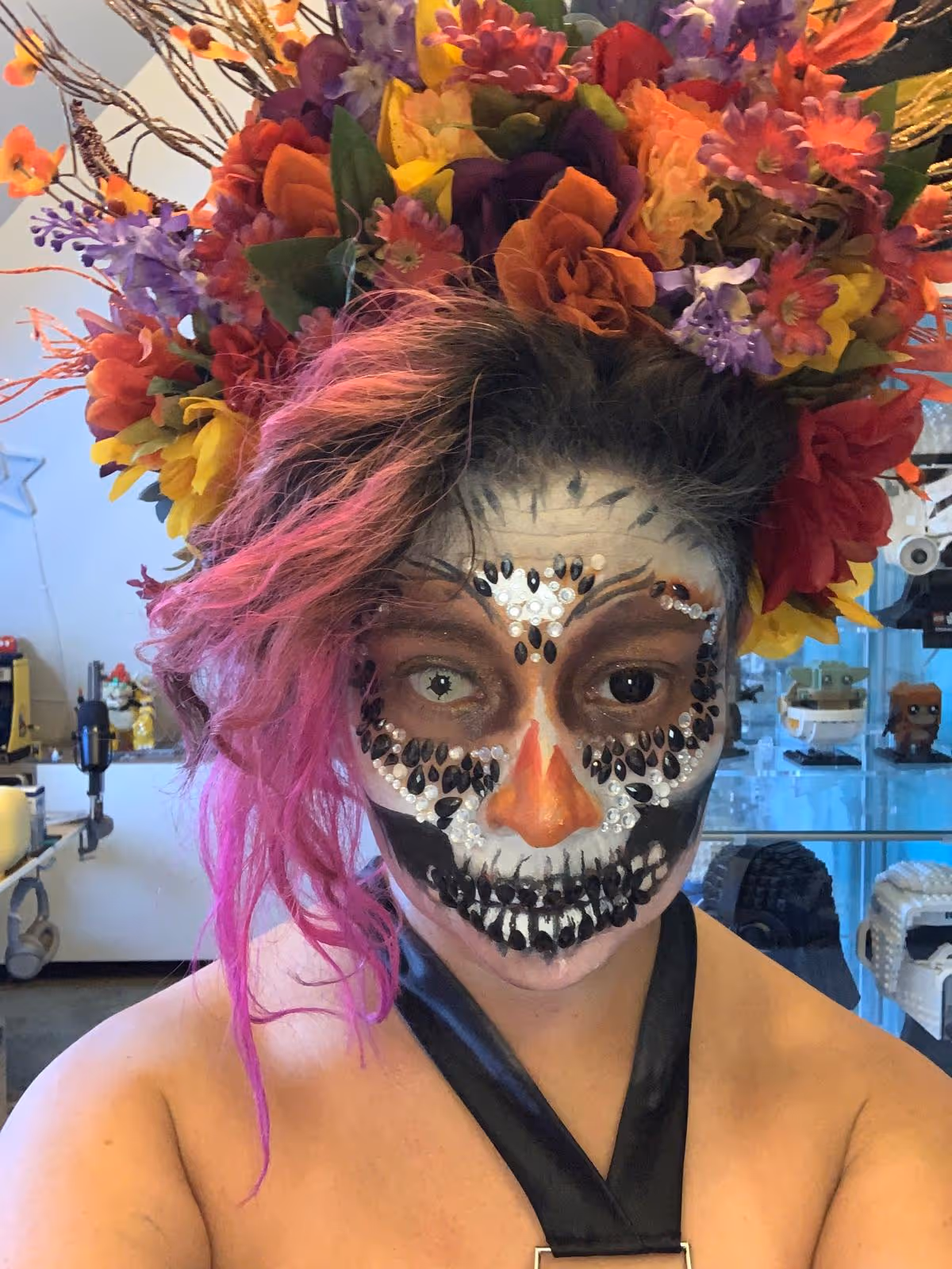 Person with skull-inspired face paint adorned with black and white jewels, bright pink hair, and a vibrant floral headpiece.