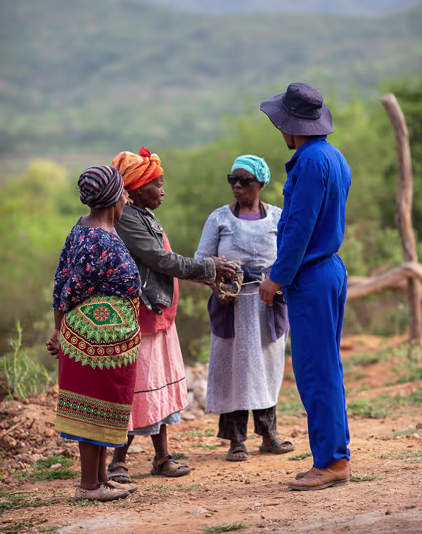 Four people standing on a dirt path outdoors, three women in colorful traditional clothing and headscarves, and a man in a blue outfit and hat, with hills in the background.