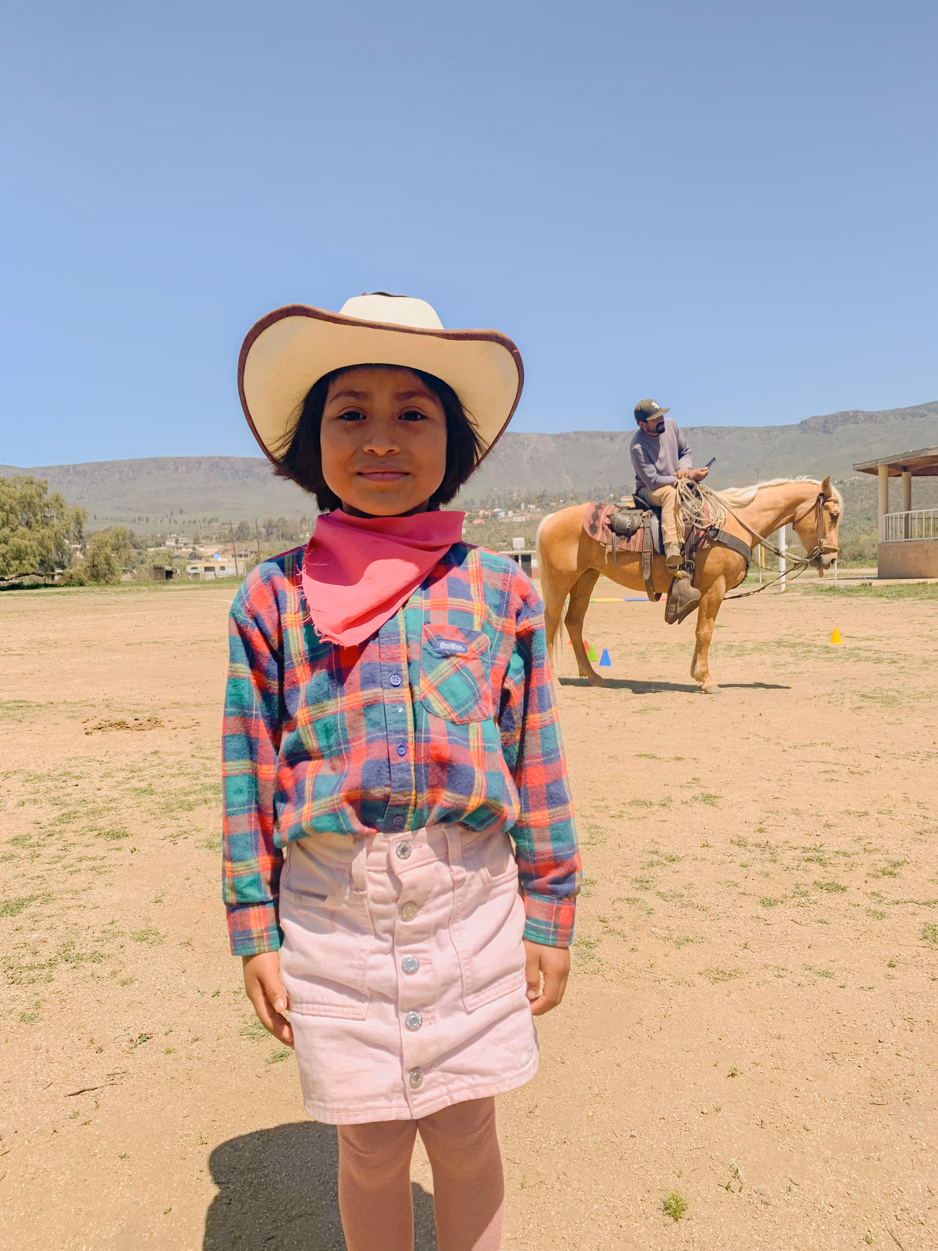Young girl wearing a cowboy hat, pink bandana, plaid shirt, and light pink skirt standing outdoors with a man on horseback in the background.