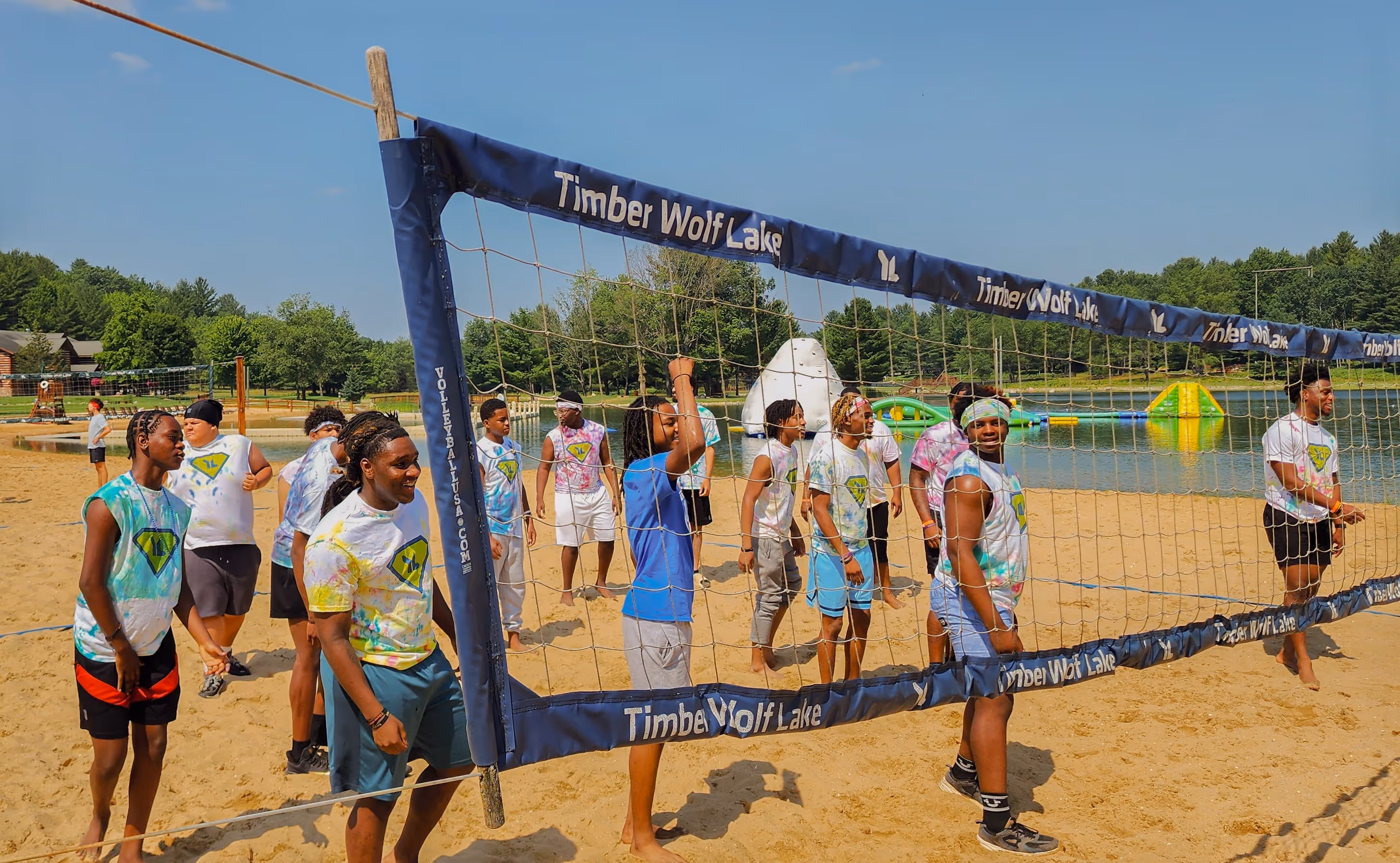 Group of young people in colorful shirts playing volleyball on a sandy beach beside a lake with inflatable water toys.