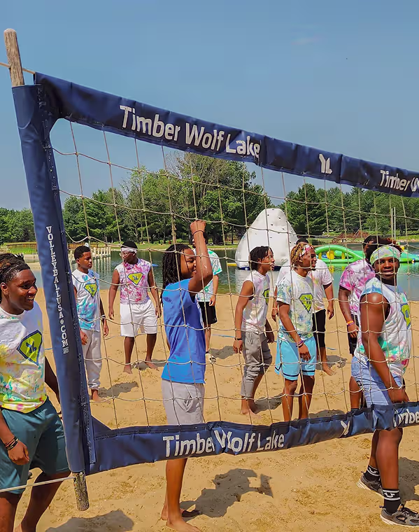 Group of young people in colorful shirts playing volleyball on a sandy beach beside a lake with inflatable water toys.