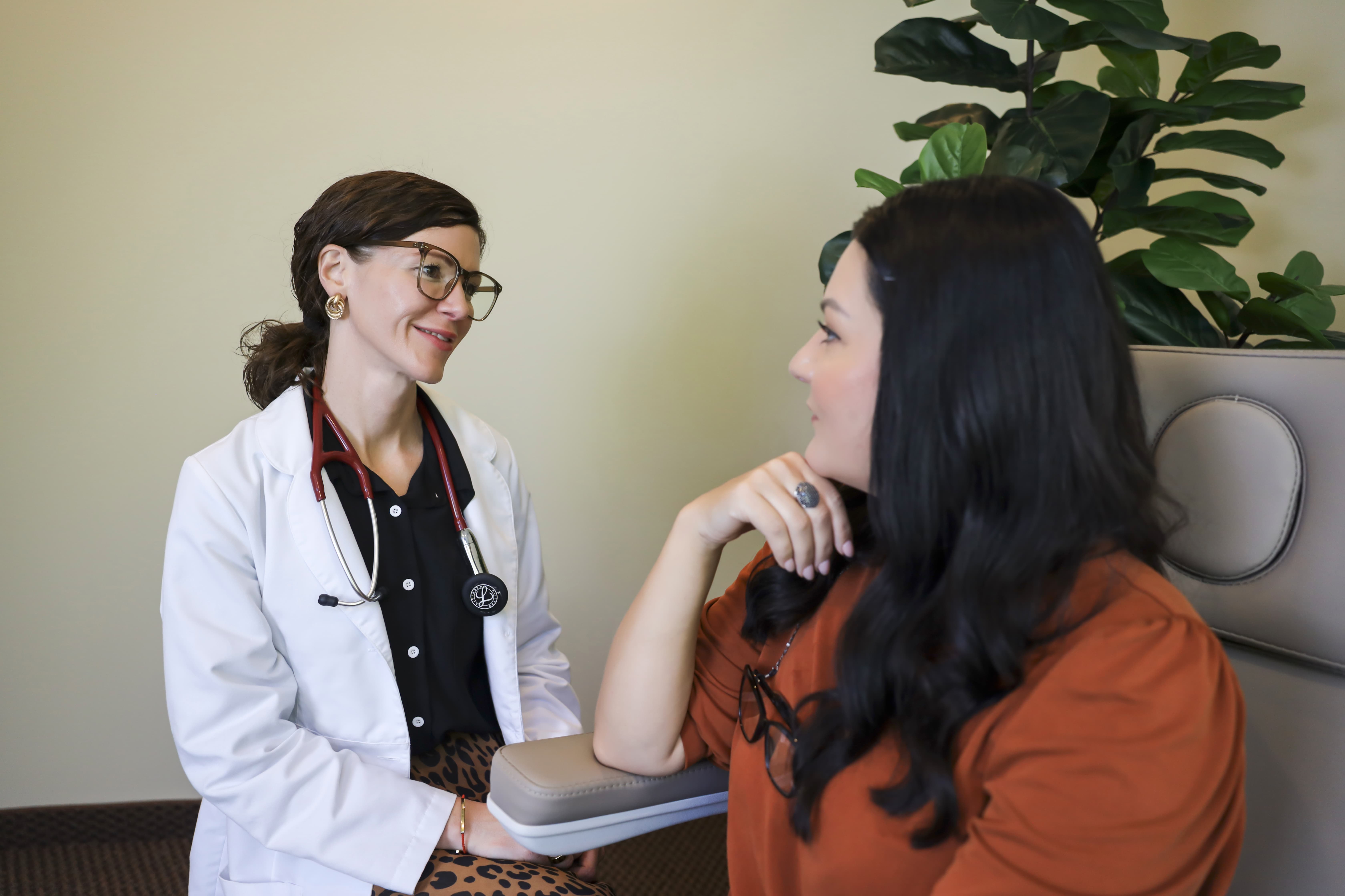 Female doctor in a white coat and stethoscope smiling and talking to a seated female patient in a consultation room.