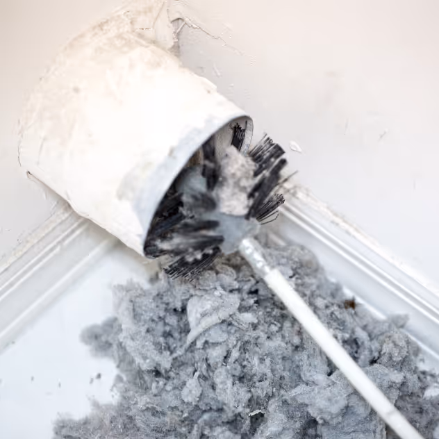 Close-up of a clogged dryer vent brush covered in lint and dust near a white wall corner.