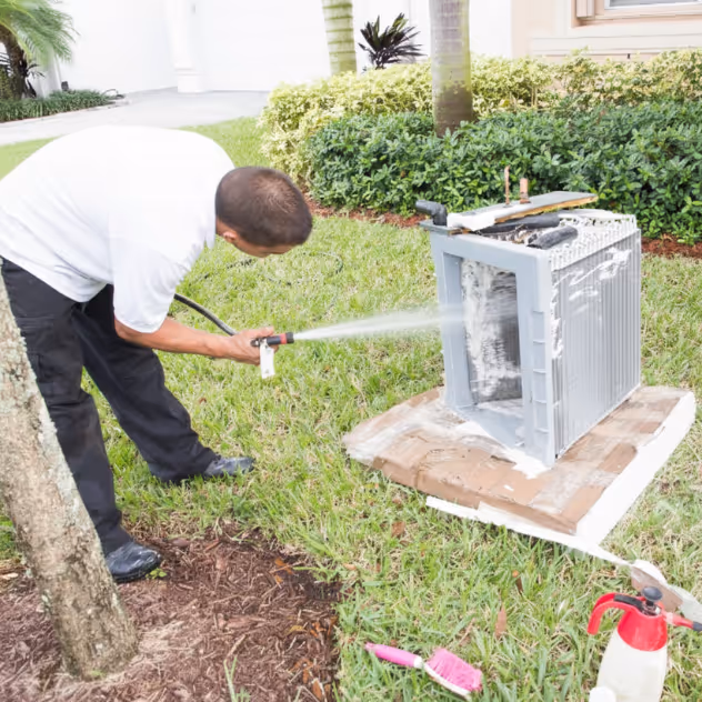 A man in a white shirt cleaning a large air conditioning coil outside with a water hose on green grass.