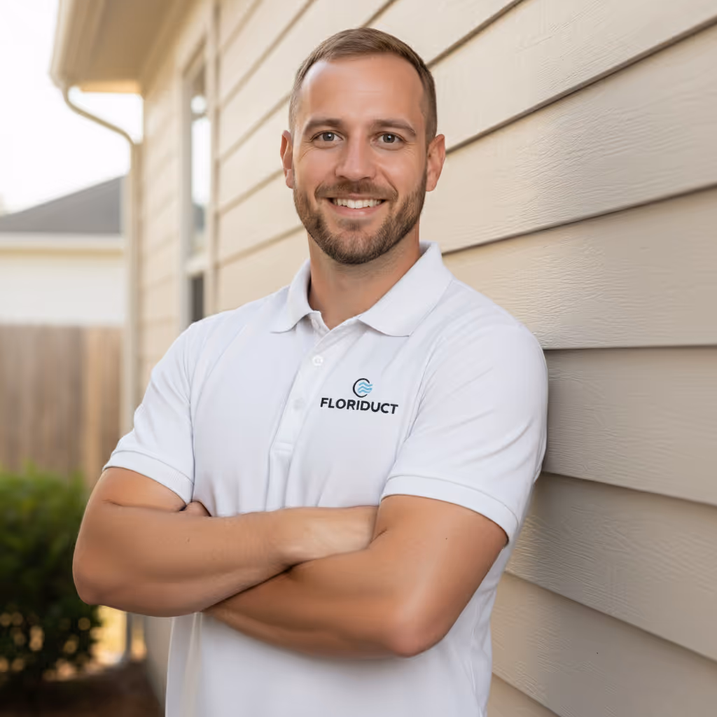 Smiling man in a white polo shirt with Floriduct logo standing with arms crossed against beige siding wall.