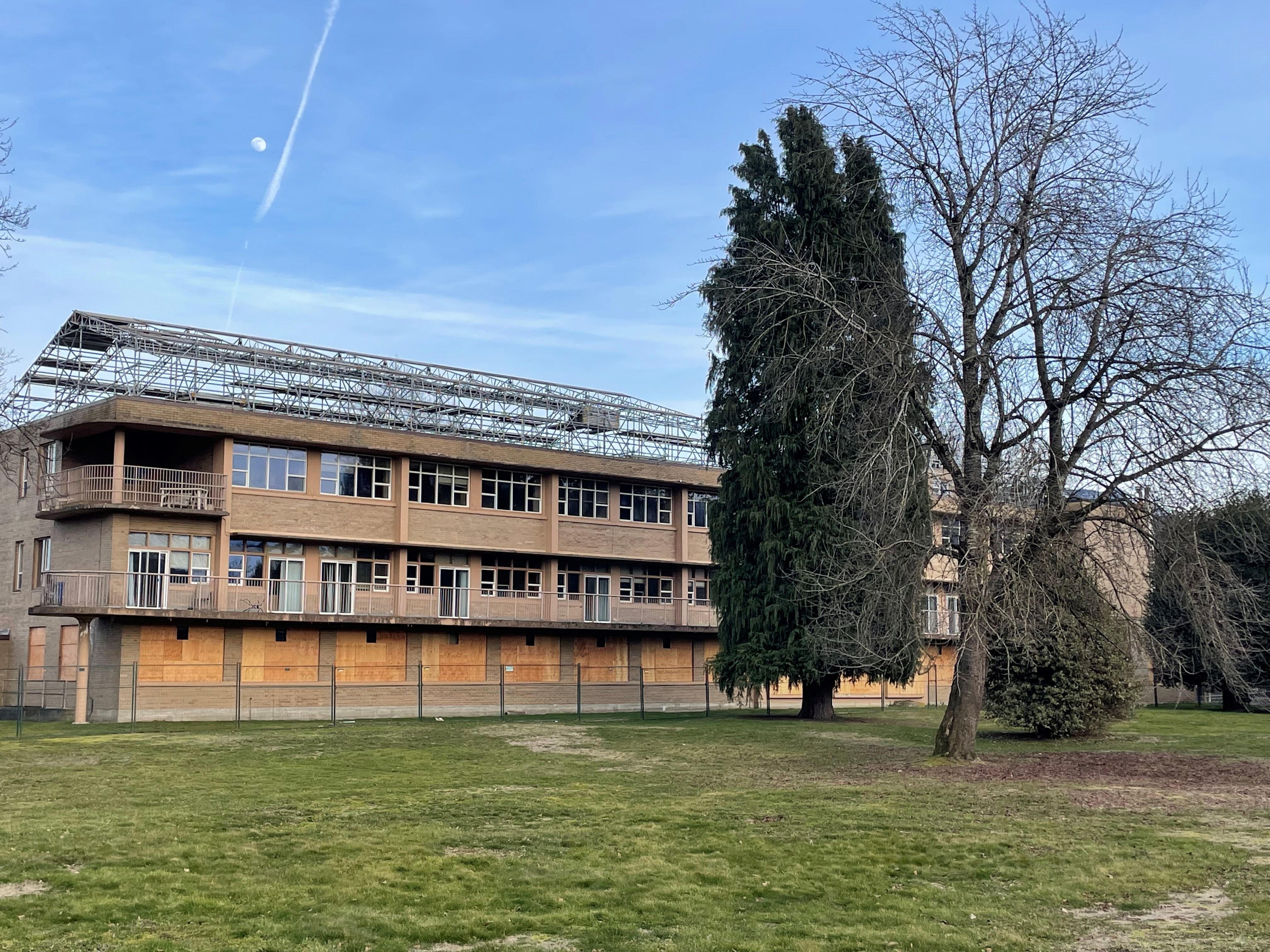 Vacant multi-storey building with boarded-up ground-floor windows and scaffolding along the roof, surrounded by open grass and trees.