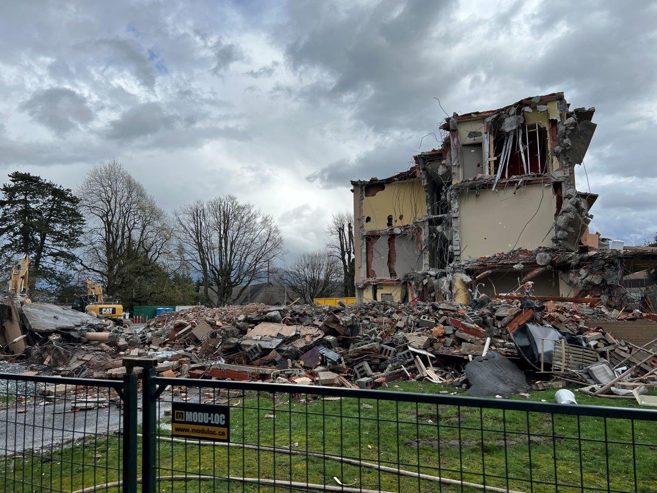 Demolition site with a heavily damaged building section standing amid large piles of rubble, machinery, and fenced-off construction zones.