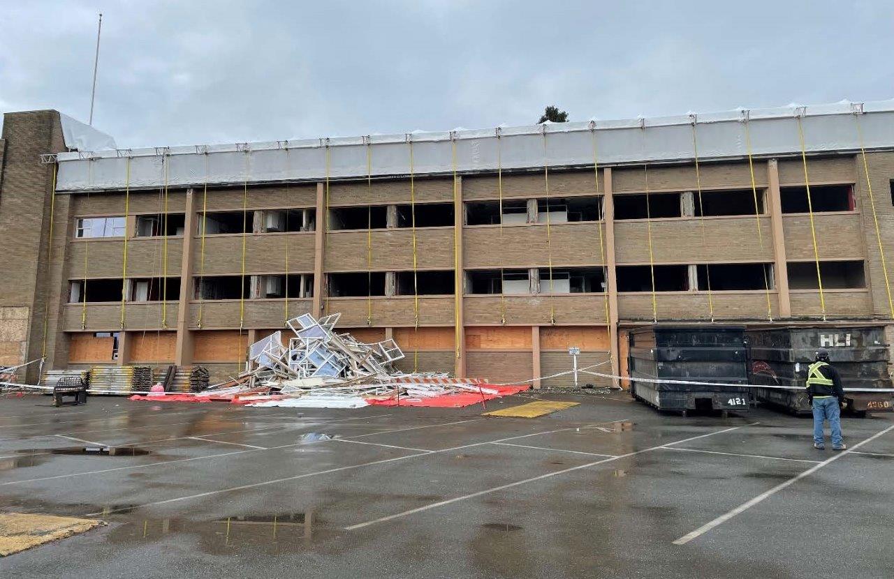 Building façade stripped of windows, with removed frames piled on the ground and disposal bins positioned in front of the structure.