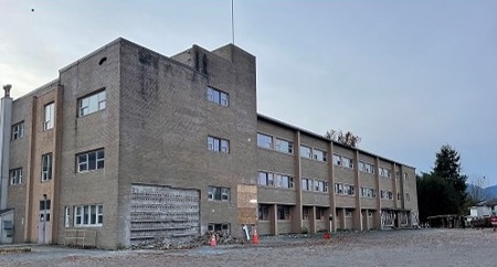 Old three-storey brick building with worn exterior walls and boarded-up entrances, standing in an empty gravel lot.
