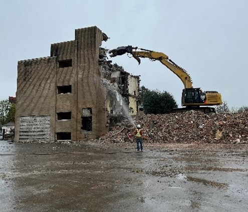 An excavator demolishes a multi-storey brick building while a worker in safety gear observes on a wet construction site.