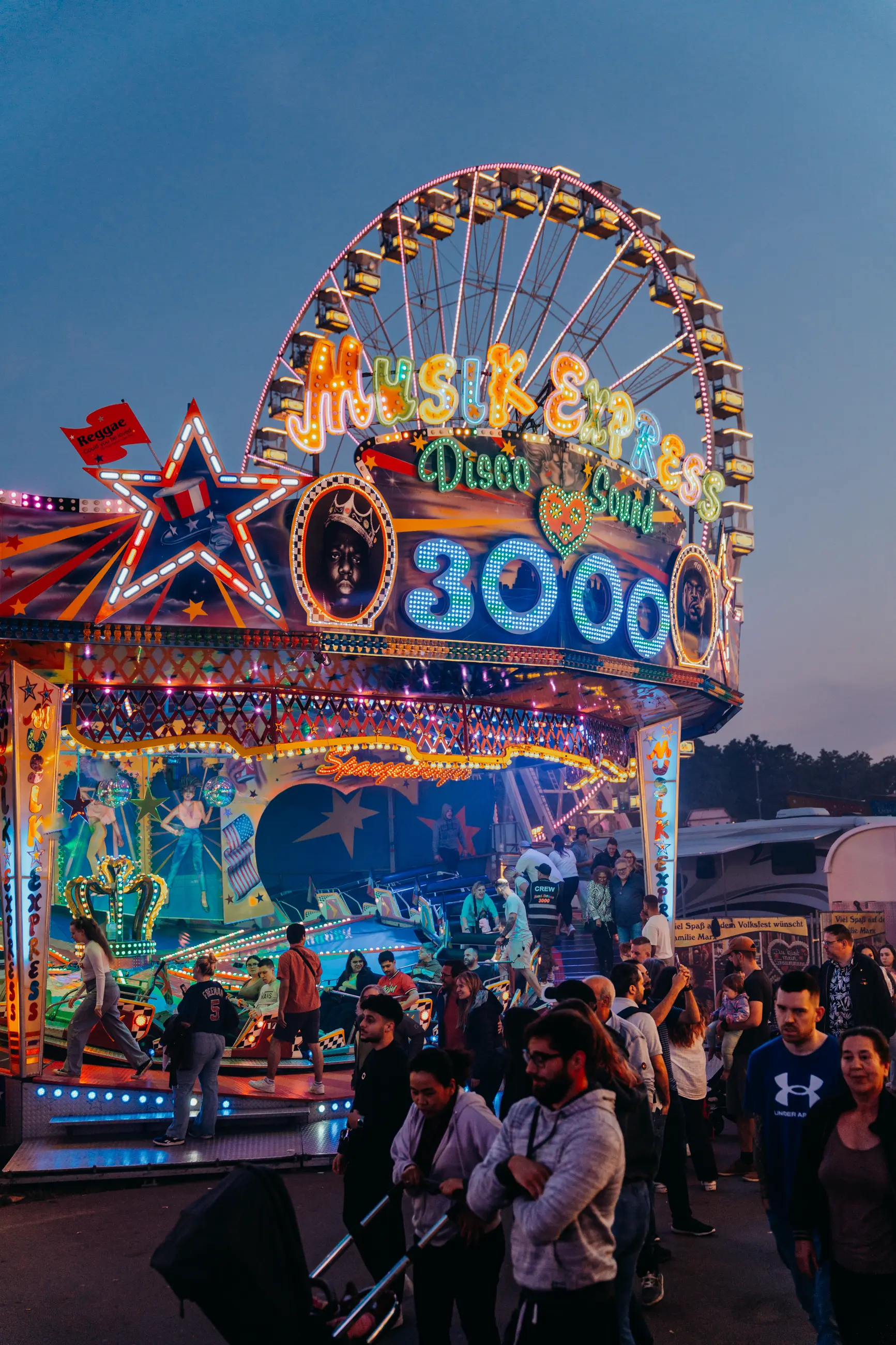 Ein illuminiertes Karussell mit der Aufschrift 'Musik Express 3000' vor einem großen Riesenrad und Menschen in einer Jahrmarktumgebung bei Dämmerung.
