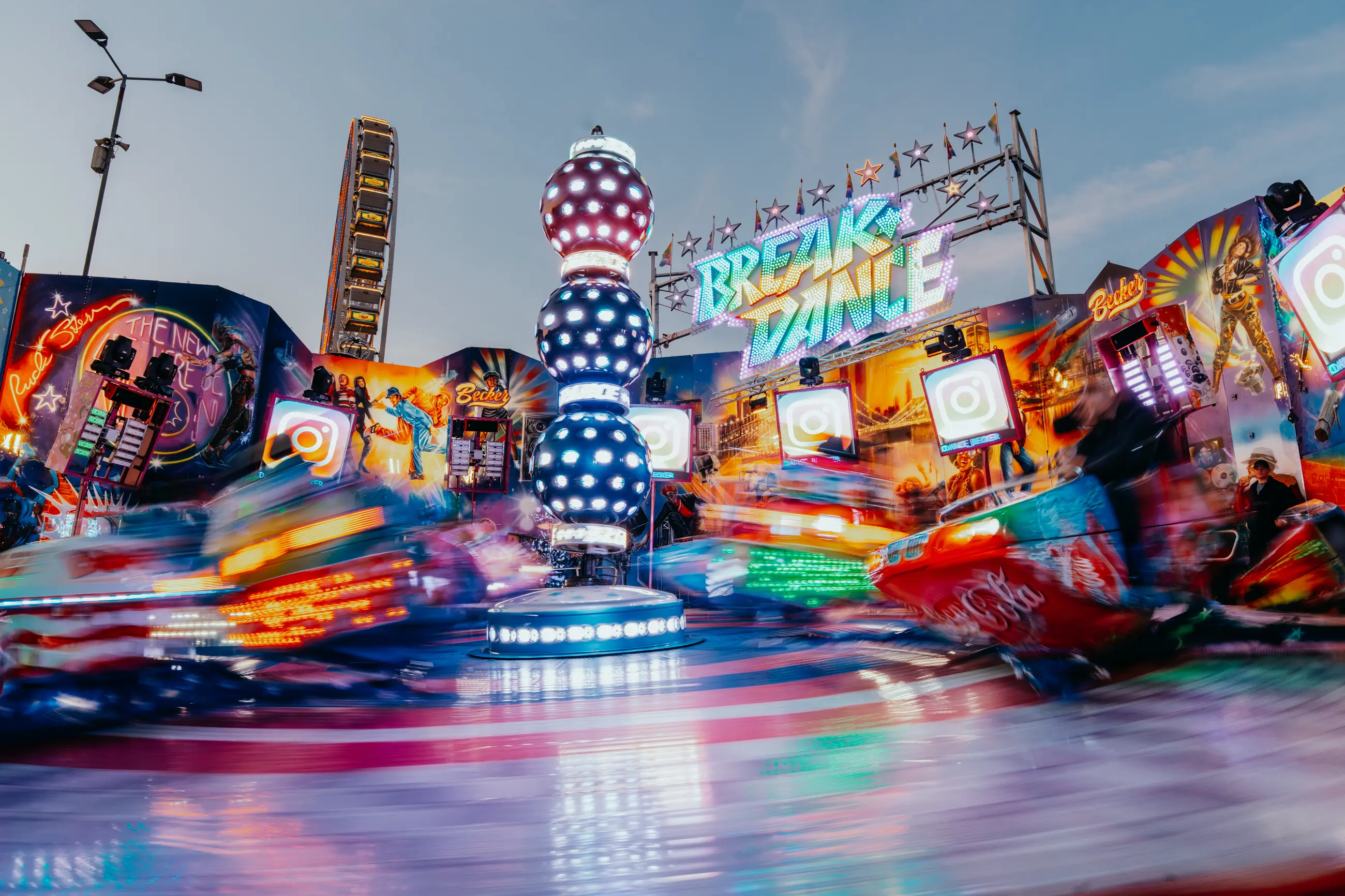 Farbige Breakdance-Fahrattraktion auf einem Jahrmarkt mit leuchtenden Lichtern und einer sich drehenden Achterbahn im Hintergrund.