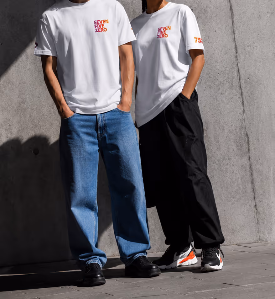 Zwei Personen tragen weiße T-Shirts mit Schriftzug SEVEN FIVE ZERO und Hände in Taschen, vor Betonwand.