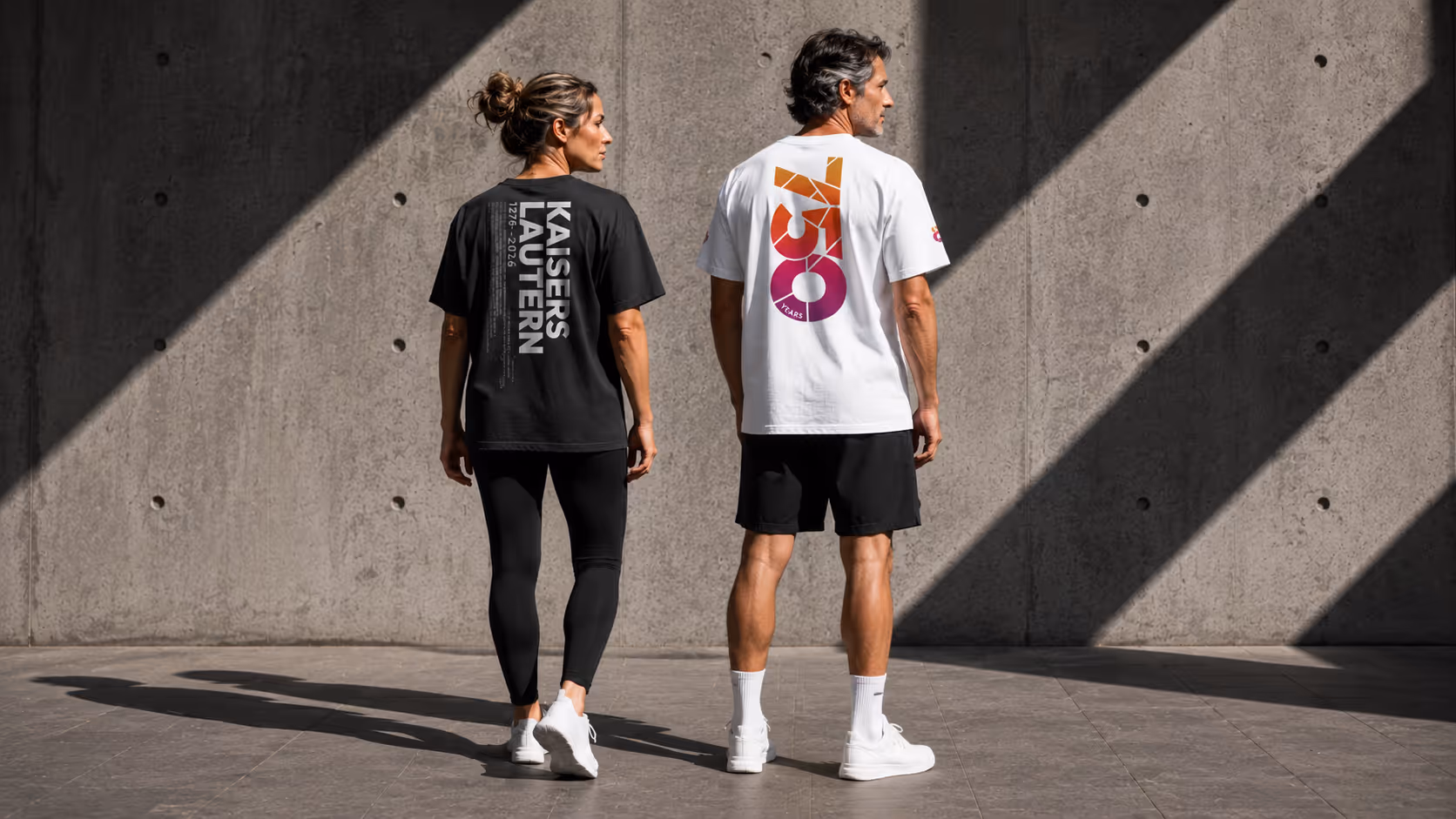 Ein Mann und eine Frau von hinten, tragen T-Shirts mit Aufdrucken vor einer Betonwand mit Lichtstreifen.