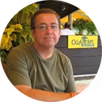 Muškarac sa naočalama u zelenoj majici sjedi ispred pozadine sa žutim cvjetovima i natpisom.